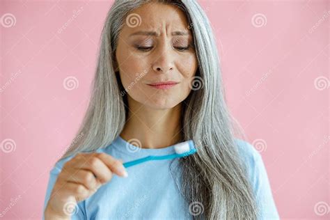 Upset Mature Asian Lady In Blue T Shirt Looks At Toothbrush Posing On Pink Background Stock