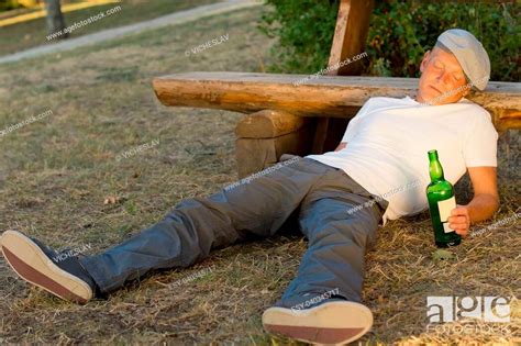 drunk man fallen asleep on the ground leaning his head on a bench holding a bottle of white wine