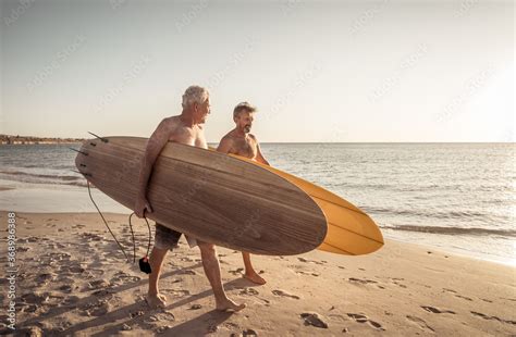 Two Senior Surfers With Surfboard Having Fun On Empty Remote Beach