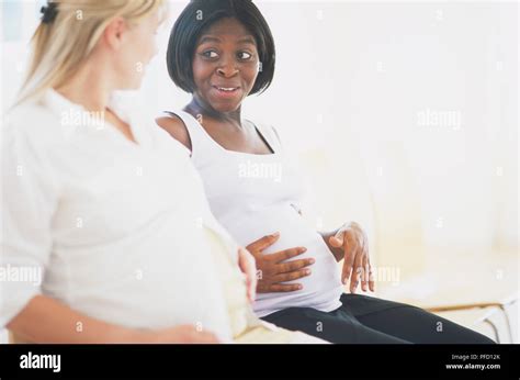 Two Pregnant Women Sitting Next To Each Other Stroking Their Belly And Chatting Side View