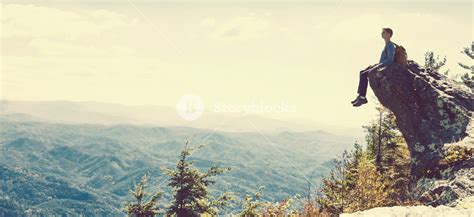 Man Sitting On The Edge Of A Cliff Overlooking The Mountains Below
