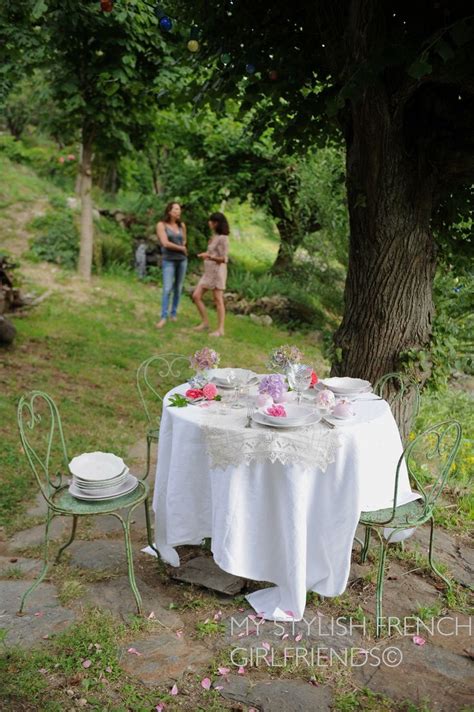 An Outdoor Table Set For Two With Plates And Cups On It Next To A Tree
