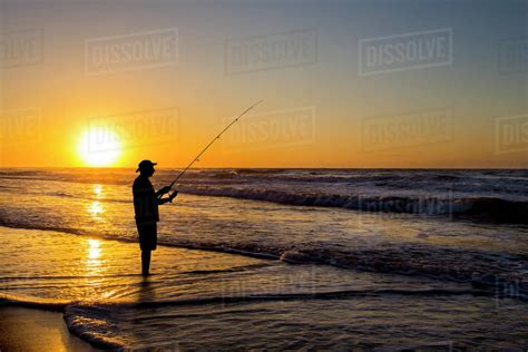 Silhouette Of Man Fishing In Waves On Beach At Sunset Stock Photo