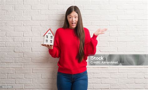 Young Chinese Real State Agent Woman Over Brick Wall Holding House Very Happy And Excited Winner