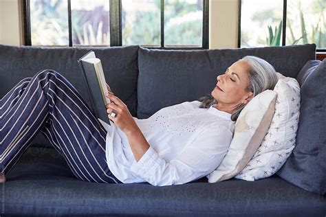 Mature Woman With Grey Hair Reading A Book Lying Down On Sofa In