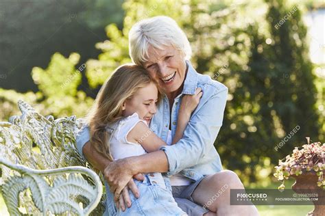 Grandmother And Granddaughter Hugging On Garden Bench Laughing Focus