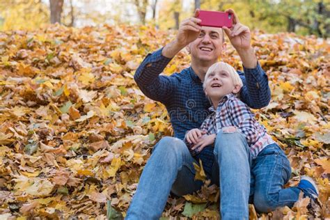 Father And Son Sitting On Autumn Leaves And Make Pictures On Phone