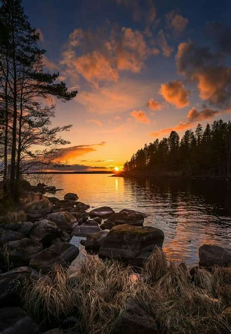 The Sun Is Setting Over A Lake With Rocks And Grass In Front Of It