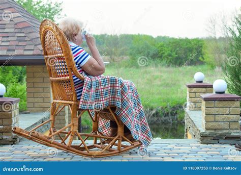 An Elderly Woman Sits In A Wicker Rocking Chair And And Drinks A Cup Of