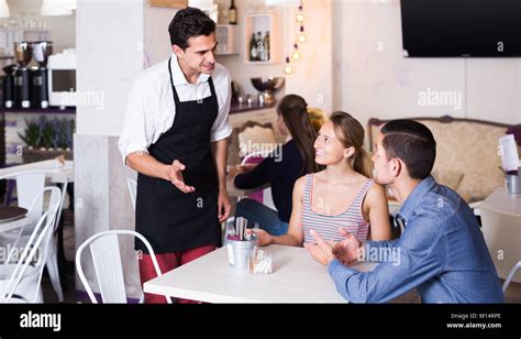 Portrait Of Young Cafe Waiter Standing At Table And Talking To Guests