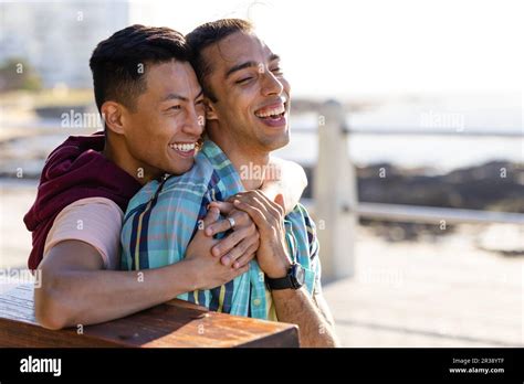 Happy Biracial Gay Male Couple Sitting On Bench And Embracing On Promenade By The Sea Stock