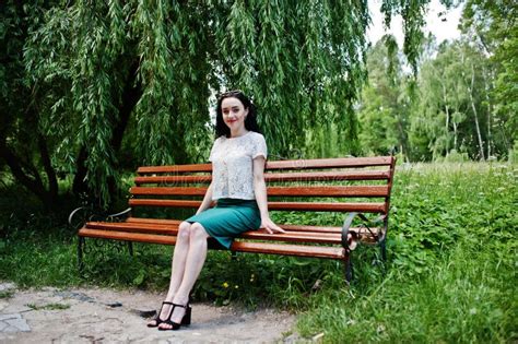 Brunette Girl In Green Skirt And White Blouse Posed At Park Sitting On