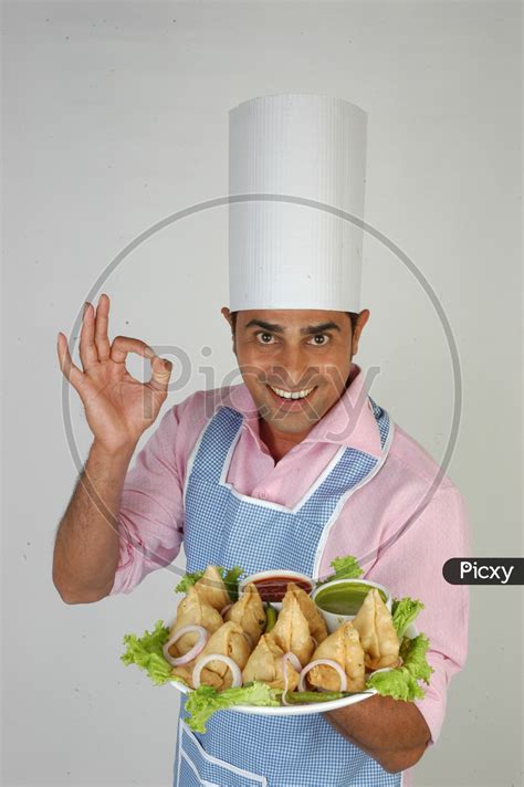 Image Of An Indian Chef In Kitchen Apron And Cap Holding Samosas Plate With An Expression On An