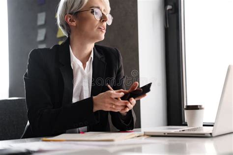 White Haired Puzzled Woman Using Mobile Phone While Working With Laptop
