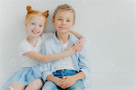 Friendly Small Girl And Boy Hug And Sit On Floor Against White Background Have Positive