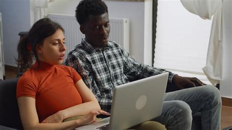 Interracial Couple Sitting On Sofa Using Laptop In Living Room Stock