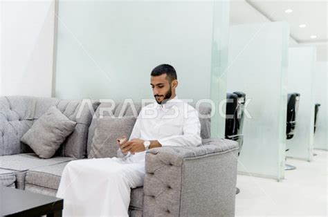 رجل سعودي خليجي جالس ينتظر دوره في صالون الحلاقة ، يقضي وقته على هاتفه المحمول ، العناية