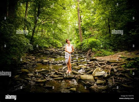 Mid Adult Woman Standing Barefoot On Rocky Riverbed Squeezing Water From Hem Of White Dress