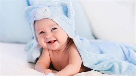 Smiling Cute Infant Is Lying Down On White Bed Covering With Blue Towel