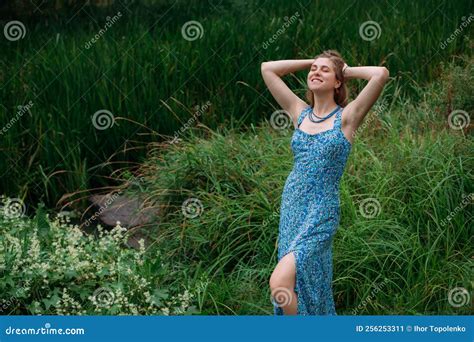 Girl With Blond Hair In A Blue Summer Dress Stands On A Green Lawn In A Forested Area And