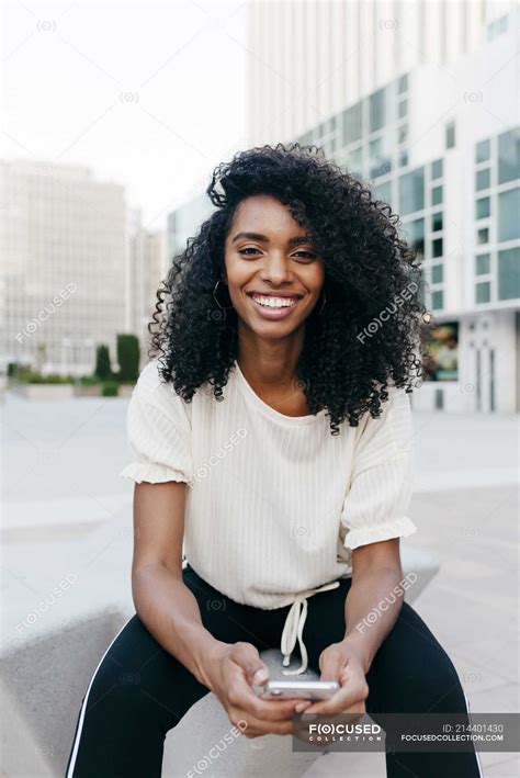 Portrait Of Smiling African American Woman Sitting On City Street With Smartphone Technology