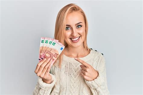 beautiful caucasian blonde woman holding 10 euro banknotes smiling happy pointing with hand and