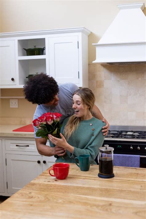 Biracial Husband Giving Roses To Cheerful Wife Having Coffee At Table In Kitchen Stock Image