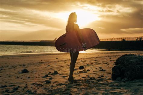 Female Surfer Posing With Surfboard On Beach At Sunset With Back Light