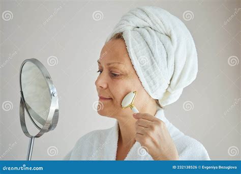 Mature Woman In White Bathrobe With Towel On Head Doing Facial Massage With Roller Looking In