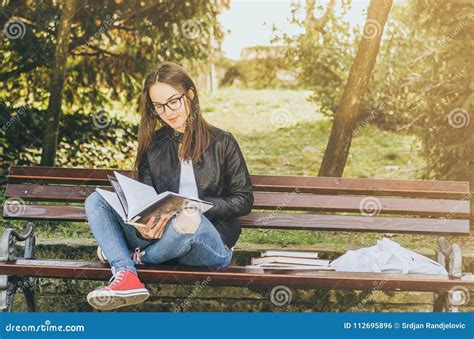 Young Beautiful School Or College Girl With Glasses Sitting On The Bench In The Park Reading The