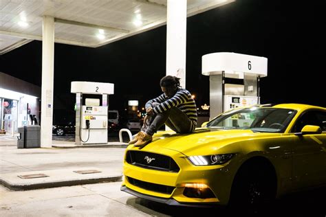 Photo Of Man Sitting On Hood Of Yellow Ford Mustang Parked At A Gas