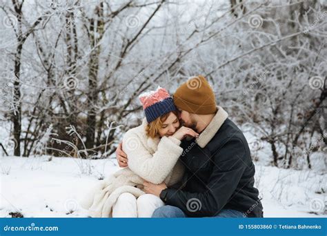 Happy Woman And Man Wearing Coats And Hats Sitting And Hugging In Winter Snow Background
