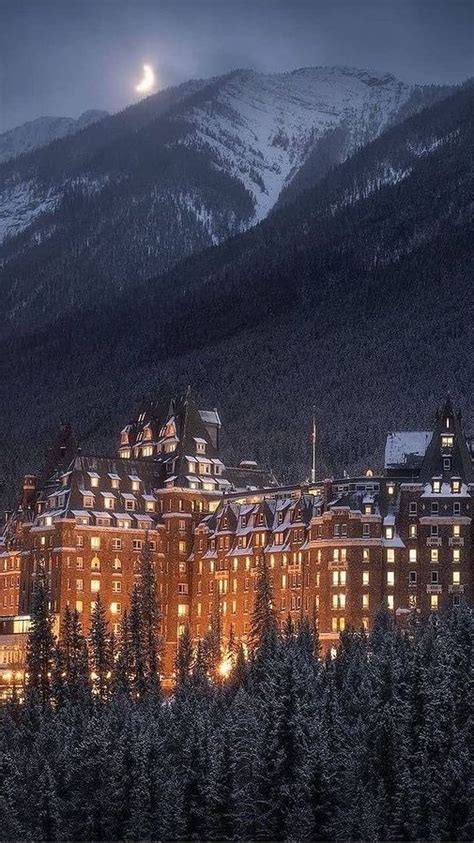 A Large Building Is Lit Up At Night In Front Of The Mountains And Snow