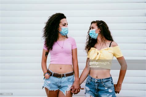 Lesbian Couple Wearing Masks Holding Hands While Standing Against Wall In City High Res Stock