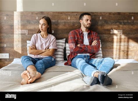 Young Frustrated Couple Sitting On Big Bed Keeping Hands And Legs