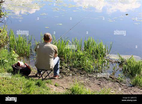 Fisherman Sitting Near The Water With A Fishing Rod Rear View Man