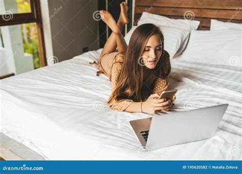 Woman Lying On Bed With Laptop Chatting In Phone In Bedroom Stock Photo