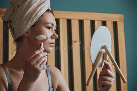Young Woman Using Jade Facial Roller For Face Massage Sitting On Bed In Bedroom Looking In The