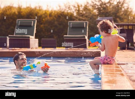 father and son squirting each other with water pistols playing in swimming pool on summer