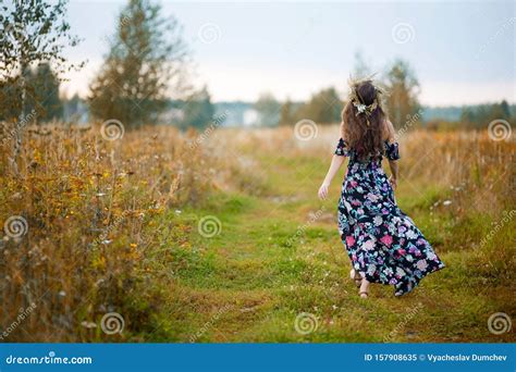 Young Girl In A Colorful Sundress With A Wreath On Her Head Goes Away Along A Country Road View