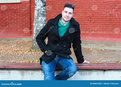 Young Handsome Man Sitting On The Bench In A Black Coat Wearing Stock