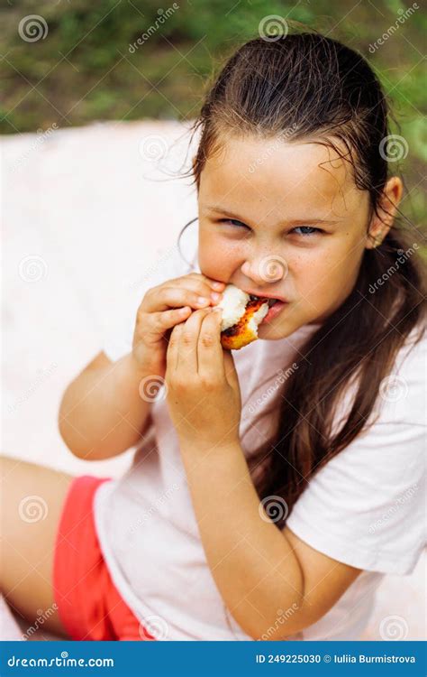 portrait of little hungry girl wth dark hair holding food in hands biting sitting in park