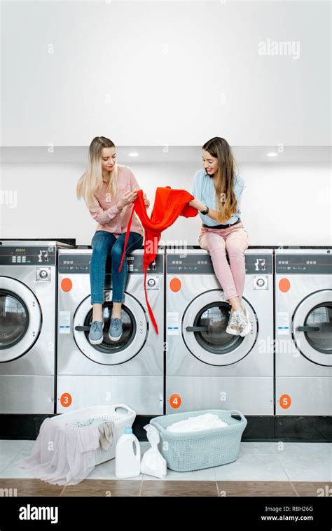 Two Cheerful Girlfriends Sorting Clothes For Washing Sitting On The Washing Machines In The