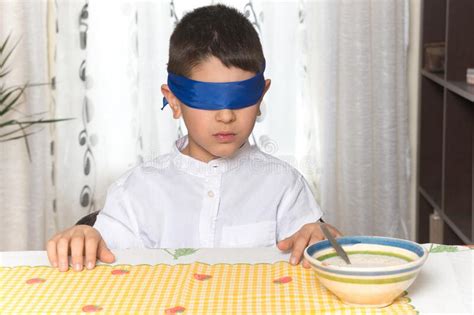 An 8 Year Old Caucasian Boy Sitting At The Table At Home Is Blindfolded