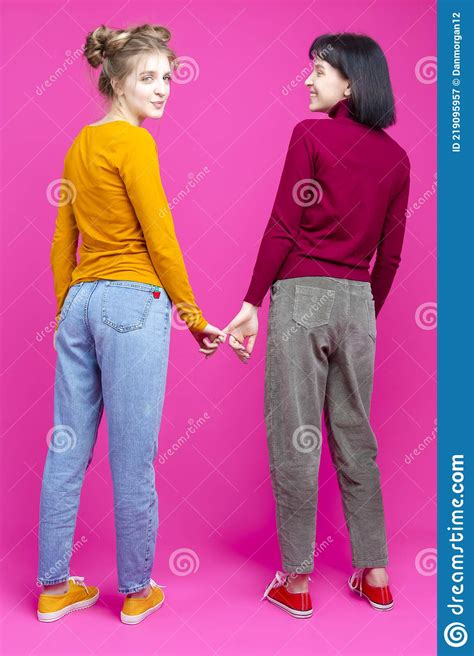 Portrait Of Youing Caucasian Lesbian Couple Posing Together With Fingers Connected Against Pink