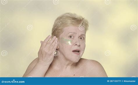 Attractive Blonde Woman Applying A Green Skin Mask Onto Her Face Under Eye Area And Looking At