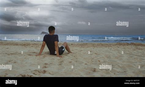 Brunette Boy Sitting On The Beach Facing The Sea With Very Cloudy Sky