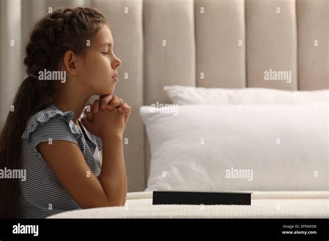Cute Little Girl Praying Over Bible In Bedroom Space For Text Stock