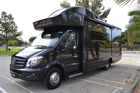 A Black Tour Bus Parked In A Parking Lot Next To Palm Trees And Grass