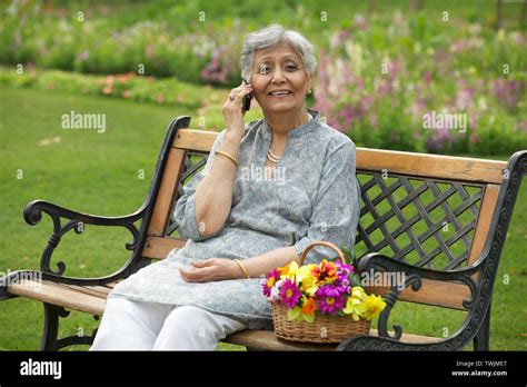 Old Woman Sitting On A Park Bench And Talking On A Mobile Phone Stock